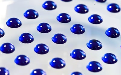 The image depicts a close-up view of numerous shiny blue water droplets scattered across a smooth white surface, each reflecting light.