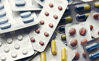 The image displays a collection of various medication pills and capsules, in multiple colors and sizes, packaged in overlapping blister packs.