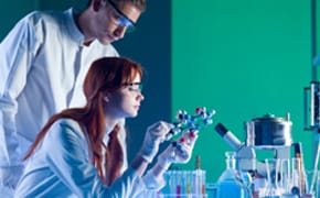 实验室里有两个穿着白大褂的人。其中一人正在仔细检查一个分子模型，另一人则在后面观察。桌子上摆放着各种实验室设备，包括试管和烧瓶，绿色背景墙形成鲜明对比。