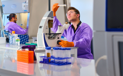 Biosafety Testing for monoclonal antibodies Lab technician in a purple coat and orange gloves using a microscope at a biosafety testing bench, with tubes and lab equipment visible in the background.