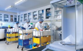 <p>Developing Robust Media Formulations</p>
Laboratory scene with scientists in white coats working at tables, surrounded by shelves of chemicals and equipment, focused on their tasks.