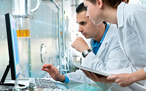 A laboratory scene depicting two scientists in white lab coats collaborating at a computer. One scientist is pointing at the screen, while the other is holding a clipboard. The background features laboratory equipment and a container with a yellow liquid, indicating an active research environment. 