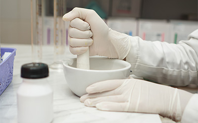 A close-up view of a gloved hand holding a pestle, grinding a substance in a white mortar bowl. A small container with a black lid is visible nearby, along with a blue basket in the background, suggesting a laboratory setting. A close-up view of a gloved hand holding a pestle, grinding a substance in a white mortar bowl. A small container with a black lid is visible nearby, along with a blue basket in the background, suggesting a laboratory setting.