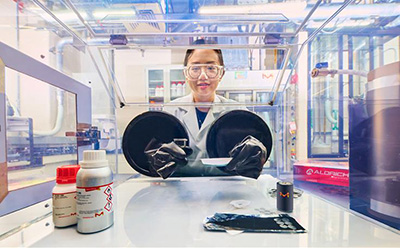 Formulation in liquid electrolyte R&D A scientist wearing safety goggles and a lab coat is working with a glove box in a high-tech laboratory. The image is taken from the outside of the glove box, showing the researcher through the transparent front panel. Various lab supplies, including chemical bottles and materials, are arranged on the bench in the foreground. The laboratory has a modern, industrial setting with advanced equipment in the background.