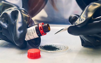 A close-up view of hands wearing black gloves pouring a black powder from a small glass bottle into a watch glass. The bottle has a red cap and a label, and the watch glass contains a small mound of the black powder A close-up view of hands wearing black gloves pouring a black powder from a small glass bottle into a watch glass. The bottle has a red cap and a label, and the watch glass contains a small mound of the black powder