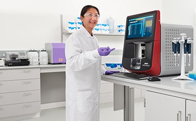 Scientist with xMAP® INTELLIFLEX® Instrument Female scientist in a lab with white coat, goggles, and gloves standing in front of a bench pointing to an xMAP® INTELLIFLEX® instrument that has pipettes and MIILIPLEX® multiplex immunoassay kits next to it.