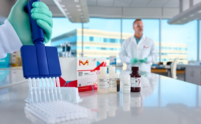 Scientist Gloved Hand Holding a Pipette Gloved hand holding a blue pipette over a lab bench with small bottles and test tubes; a scientist in a white coat stands in the blurred background in a modern laboratory.