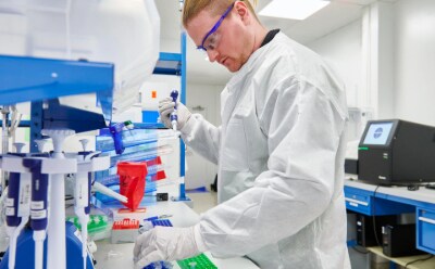 Scientist in lab coat with gloves holding a pipette by a bench