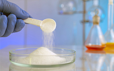 Carbohydrate powder Gloved hand scooping white carbohydrate powder into a petri dish with laboratory glassware in the background.