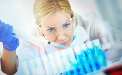 Mid 30's blond female chemist carefully measuring chemicals in a laboratory. Wearing protective mask and eyeglasses. She's using pipette to accurately put blue chemical into flasks..