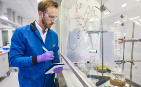 A laboratory technician in a blue lab coat and purple gloves is taking notes in front of a fume hood. Various glassware, including a round-bottom flask and a separating funnel, is visible on the counter, along with chemical structures and diagrams on a whiteboard in the background.