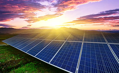 Ground-mounted solar panels capturing sunlight at sunrise, with colourful clouds and distant mountains in the background.