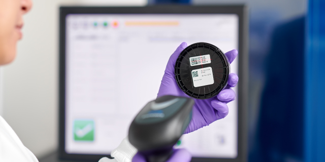 <p>From Manual to Automated QC</p>
Purple-gloved hand holds a labeled black culture plate while a handheld scanner is positioned in front of a computer screen.