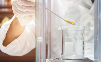 A person wearing a white lab glove is holding a spatula with a yellow substance over a glass beaker inside a laboratory fume hood.