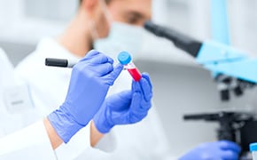 Laboratory labels A person in a white lab coat stands in a laboratory facing away from the camera, holding a clipboard and writing notes. The laboratory is equipped with shelves stocked with various lab equipment such as beakers, flasks, test tubes, and bottles containing chemicals labeled with different colors.