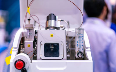Mass Spectrometer A detailed view of a protein mass spectrometry machine, featuring various components such as tubes, wires, and dials, with a visible emergency stop button and cautionary yellow label. In the background, a scientist in a lab coat works within a laboratory setting, highlighting the machine’s use in scientific research and technological advancement.