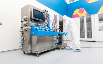 A technician in a white protective suit and blue shoe covers is operating a large, stainless steel pharmaceutical machine with blue accents, which features various tubes and a digital screen. The background shows a brightly colored, modern laboratory environment with geometric patterns on the walls.