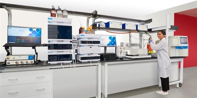 <p>Enable glycosylation monitoring during perfusion cell culture</p>
A laboratory setting featuring advanced scientific equipment, including multiple chromatography systems and a computer monitor displaying data. A researcher in a lab coat is actively engaged in an experiment at a workbench, surrounded by various instruments and supplies essential for laboratory analysis.