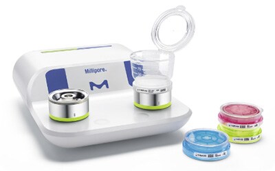 密理博 Milliflex Oasis® 过滤系统，其设计简洁大方：白色底座上放置着两个圆形过滤单元，顶部配有一个透明容器，周围摆放着几个蓝色和粉色的培养皿，所有元素均陈列于洁净的白色背景之上。