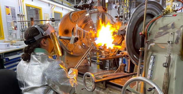 Glass Shop Glassblower 圆底烧瓶位于车床中,玻璃技师将火焰引至烧瓶颈部。玻璃技师身着防护服,包括专用外套、手套和面罩。