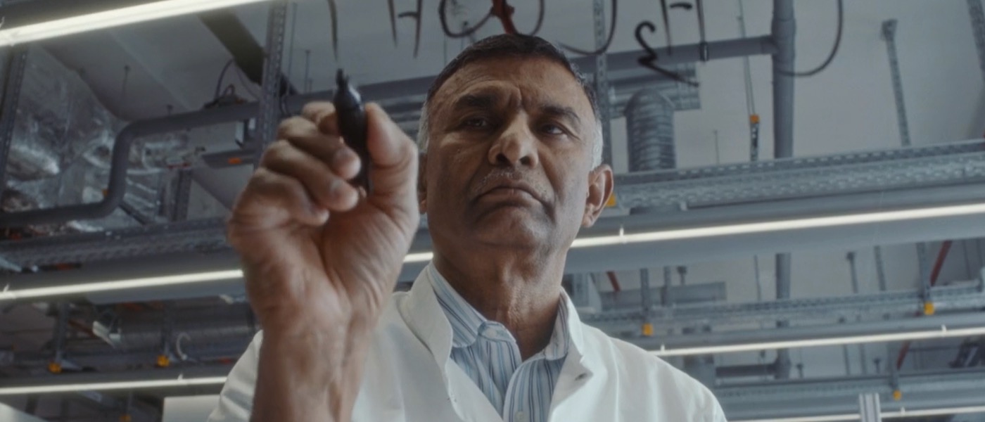ponnusamy-green-chemistry Dr. Ponnusamy wears a lab coat is writes out chemical compounds on glass with a marker.