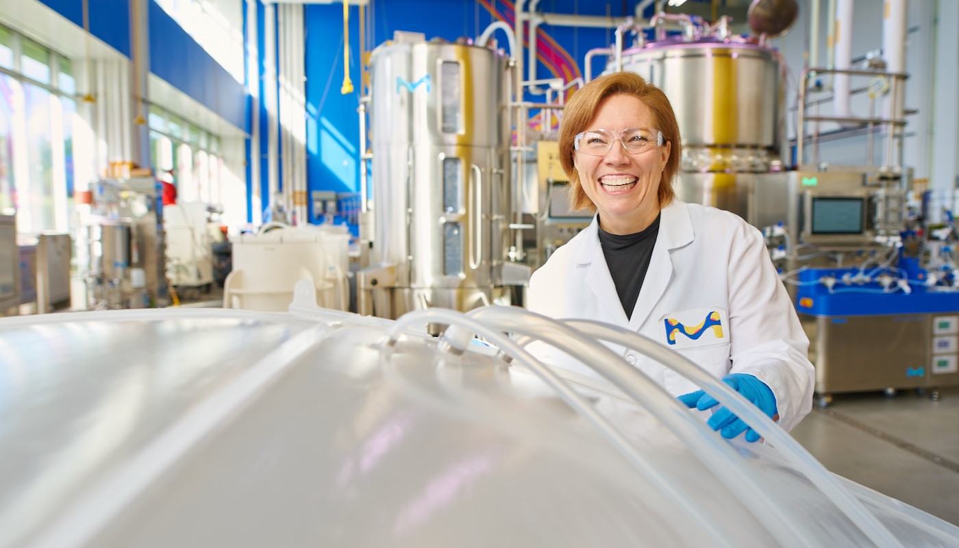 Shannon Cleveland Holds Ultimus Bag Shannon 正穿着实验室外套、戴着蓝色手套和护目镜,站在M Lab(非 GMP 生物制造环境)中。她正微笑地拿着一只垃圾袋(左下角)。