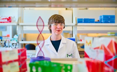 Be the Base Edit Boss A laboratory setting featuring a person in a white lab coat standing in front of shelves filled with various laboratory supplies. In the foreground, there is a model of a DNA double helix, and colorful boxes are visible, indicating different products or samples.