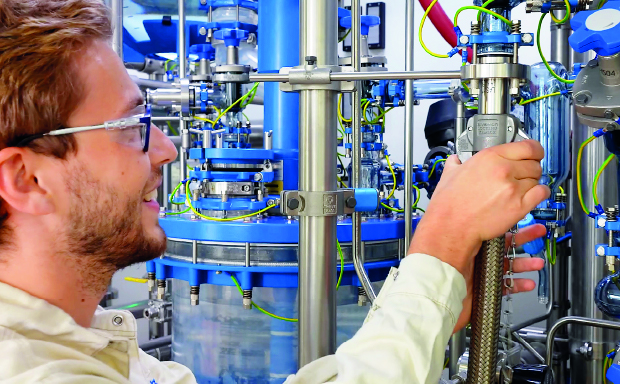 A male production worker in the Schaffhausen pilot plant facilities is connecting a solvent hose to the reactor system, preparing for upcoming manufacturing activities.