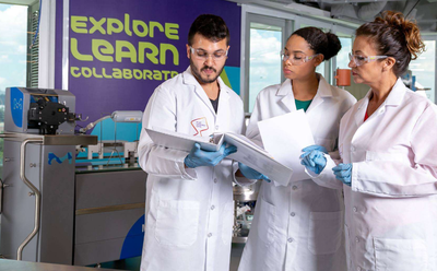 Life Cycle Assessment Three scientists in white lab coats review documents beside laboratory equipment; a wall graphic reads “Explore, Learn, Collaborate.”