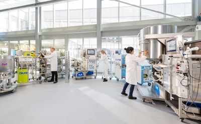 Particulate risk mitigation in single-use systems A pharmaceutical production suite with several scientists monitoring and operating stainless-steel bioprocessing systems, tubing, and control units in a spacious, light-filled facility.