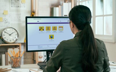 mConfig™ Configurator Home Screen A woman with dark hair in a ponytail sits at a desk, looking at a computer monitor displaying a colorful dashboard with six yellow tiles.