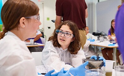 Curiosity Programming Two children are engaged in a hands-on science activity, wearing protective lab coats and gloves. One child, holding a container, is demonstrating something to another child who is attentively listening, while a few adults and other participants are visible in the background, creating an interactive and educational environment.