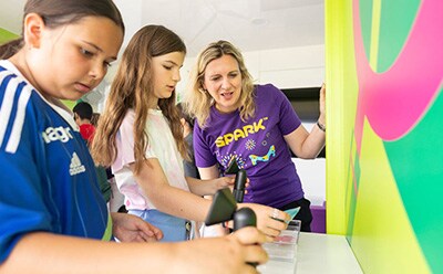 SPARK™ program Three children are engaged in an interactive activity at a colorful educational booth, with one child holding a device while another observes intently. An adult, wearing a purple SPARK™ volunteer shirt, is guiding them through the activity, promoting hands-on learning and engagement. The vibrant background features bright colors and patterns, enhancing the lively atmosphere of the event.