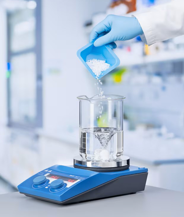 emsure-granulated-salts-dissolution-in-beaker Gloved hand transferring a white powder sample into a beaker placed on a digital analytical balance in a laboratory setting.
