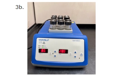 Glass vials in a digital two-block heater A digital heat block device labeled as "3b." The heat block is blue with a white control panel and a digital display. The display shows a temperature setting of 80°C and a time setting of 20 minutes. On top of the heat block, there are eight glass vials, each capped with a black screw cap. The vials are placed in a metallic block that holds them securely. The device is positioned on a black surface with a light-colored background.