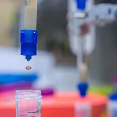 ): Photograph of a protein solution eluting from a resin column, with a drop of solution falling into a clear conical tube, in a laboratory setting.