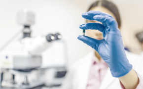 Pharma Applications Carbohydrates Close-up of a gloved hand holding a transparent red pill, with a blurred woman’s face examining it in the background with a microscope indicating a clinical or scientific setting.