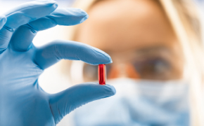 Pharma Applications in Carbohydrates Close-up of a gloved hand holding a transparent red pill, with a blurred woman’s face examining it in the background indicating a clinical or scientific setting.