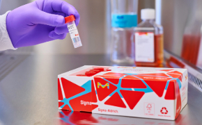 A gloved hand reaches into the tissue culture hood from the top left side of the image. It is holding a MilliShot vial that it has removed from the open box of product in the foreground. The box has vials in it, with the caps visible. In the background, on the right, is a bottle of cell culture media next to a Stericup filter.