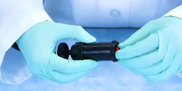 <p>Millipore<sup>®</sup> 用于石棉空气监测的气溶胶监测仪</p> A man in blue golves handling a black instrument in lab environment