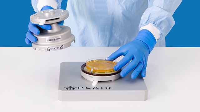<p>Rapid-C+ Air Monitoring System</p>
Blue-gloved hands position a culture plate on a P L A I R air-monitoring device while holding its sampling component.