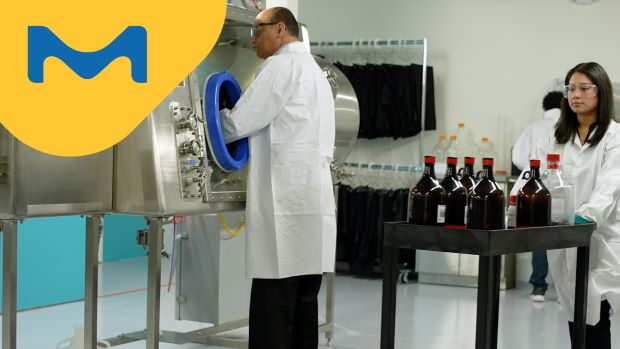 <p>The Secret of HPAPI Production Success</p>
A male scientist in a lab coat is using an isolator, while a colleague female scientist in a lab coat is transferring chemicals into the training facility behind him.