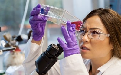 身着白大褂、戴着紫色手套的科学家手持并检查一个装有红色液体的透明小瓶。可见实验室设备，包括显微镜的一部分，表明这是一个科学实验室环境