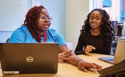 Two individuals engaged in a discussion while working on laptops at a table, background includes an office or co-working space with light blue walls and windows