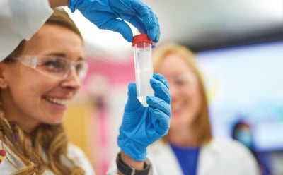 A person in blue gloves holding a test tube filled with a clear liquid, another person nearby in a laboratory setting. Background includes various laboratory equipment and bright lighting