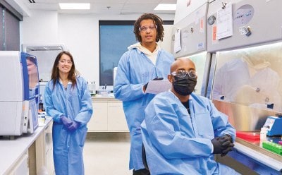 Three lab-coated individuals collaborating in a well-organized laboratory, background includes white cabinets, stocked shelves, and informational posters