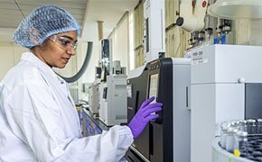 A female scientist wearing a white lab coat, a blue hairnet, safety goggles, and purple gloves is operating a sophisticated analytical instrument in a well-equipped laboratory. She is standing next to a black and silver machine with a touchscreen interface, carefully pressing the screen to adjust settings. The laboratory is filled with high-tech equipment, including a white gas chromatograph system with a series of sample vials neatly arranged in a circular tray. The background features an organized workspace with metallic and white surfaces, pipes, and analytical instruments mounted on the walls. A female scientist wearing a white lab coat, a blue hairnet, safety goggles, and purple gloves is operating a sophisticated analytical instrument in a well-equipped laboratory. She is standing next to a black and silver machine with a touchscreen interface, carefully pressing the screen to adjust settings. The laboratory is filled with high-tech equipment, including a white gas chromatograph system with a series of sample vials neatly arranged in a circular tray. The background features an organized workspace with metallic and white surfaces, pipes, and analytical instruments mounted on the walls.