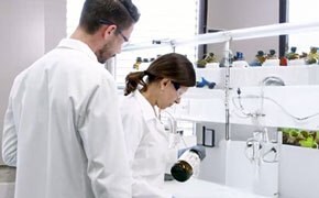 In a bright and modern laboratory, two scientists wearing white lab coats and safety goggles are engaged in an analytical procedure. The female scientist in the foreground, with dark brown hair tied back, is carefully holding a dark amber glass bottle with a black cap, tilting it slightly as she examines its contents. The male scientist standing beside her, with short brown hair and a beard, observes her actions attentively. The background features a well-organized laboratory bench with white shelves lined with multiple brown glass reagent bottles, some with blue and yellow caps. Stainless steel laboratory fittings, including a faucet and tubing system, are mounted on the wall. Large windows allow ample natural light to illuminate the workspace, emphasizing a clean, sterile environment. In a bright and modern laboratory, two scientists wearing white lab coats and safety goggles are engaged in an analytical procedure. The female scientist in the foreground, with dark brown hair tied back, is carefully holding a dark amber glass bottle with a black cap, tilting it slightly as she examines its contents. The male scientist standing beside her, with short brown hair and a beard, observes her actions attentively. The background features a well-organized laboratory bench with white shelves lined with multiple brown glass reagent bottles, some with blue and yellow caps. Stainless steel laboratory fittings, including a faucet and tubing system, are mounted on the wall. Large windows allow ample natural light to illuminate the workspace, emphasizing a clean, sterile environment.