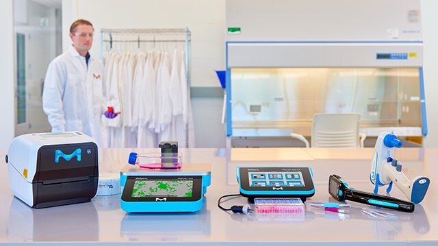 Connected Lab Instrumentation Five instruments sitting on a laboratory bench with a scientist in a lab coat walking past in the background.