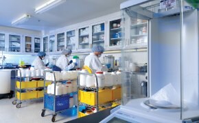 Laboratory scene with scientists in white coats working at tables, surrounded by shelves of chemicals and equipment, focused on their tasks.